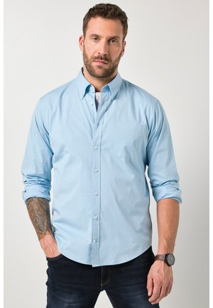 Man with beard wearing a light blue button-up shirt with rolled-up sleeves, dark jeans, a wristwatch, and a tattoo on his left forearm.