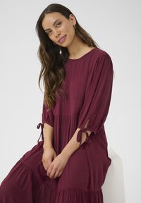 Burgundy klänning med lagerdesign, trekvartsärm och knytdetaljer vid ärmsluten. Tyget verkar texturerat och lätt i vikt.