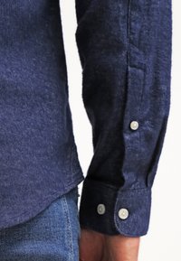 Manche et poignet d’une chemise en denim bleu foncé avec des boutons blancs, portée sur un jean bleu sur le bras et la taille d’une personne.