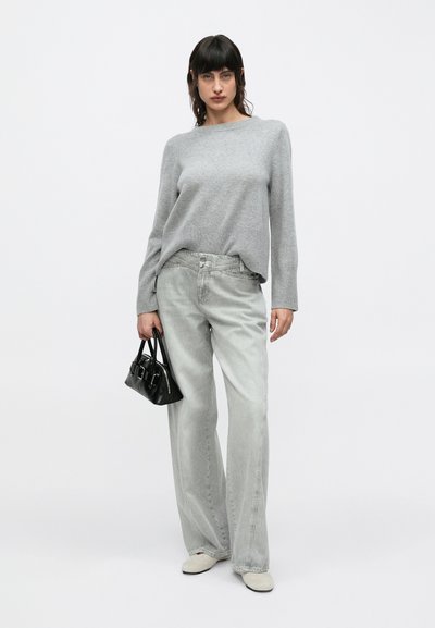 Γκρι πουλόβερ με μακριά μανίκια, ανοιχτό γκρι παντελόνι καμπάνα, και μικρή μαύρη τσάντα χειρός. Παπούτσια σε ανοιχτό χρώμα. Μινιμαλιστικός σχεδιασμός.