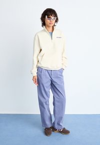 Sweatshirt zippé crème avec une poche kangourou, associé à un pantalon large violet clair et des chaussures brunes. Modèle portant des lunettes de soleil miroitées.