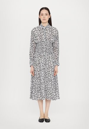 Robe à manches longues avec un motif floral noir et blanc, taille smockée et col, atteignant sous le genou. Chaussures plates noires.