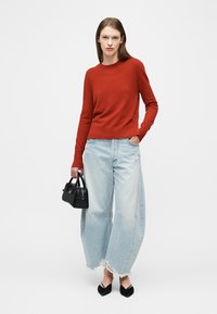 Femme portant un pull rouge à manches longues, un jean large bleu clair déchiré, des ballerines noires à bout pointu et tenant un petit sac à main noir.