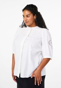 Vit kortärmad blus med volang framtill och spetsdetaljer på ärmarna. Tillverkad av lättviktig tyg, med avslappnad passform och rundad halsringning.