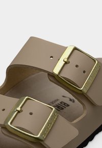 Ανοιχτό μπεζ σανδάλια με δύο ρυθμιζόμενες αγκράφες σε μεταλλικό χρυσό χρώμα, με υφή στην ενδιάμεση σόλα και λείο ανώτερο υλικό.