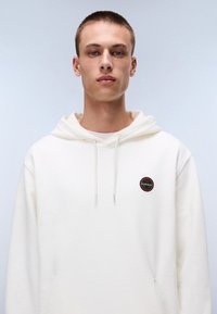 Hvid hoodie lavet af blødt bomuld, med en rund logo-patch på venstre bryst, snørelukning i hætten og en kængurulomme.