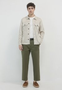 Beige Hemdjacke mit zwei Brusttaschen, schwarzen Knöpfen, olivgrünen geraden Hosen und braunen Schnürschuhen. Einfaches, sauberes Design.