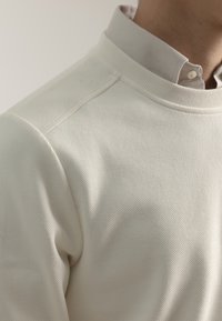 Suéter de color crema con canalé que presenta un alto cuello de canalé y una camisa con cuello en un tono gris claro debajo.