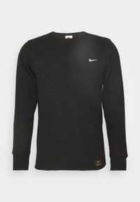Pull noir à manches longues, texture côtelée, col rond, avec le logo Nike blanc sur la poitrine. Présente une petite étiquette de marque à l'ourlet.