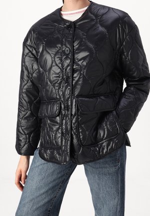 Personne portant une veste matelassée noire boutonnée avec des poches à rabat avant et un jean bleu, debout devant un fond uni.