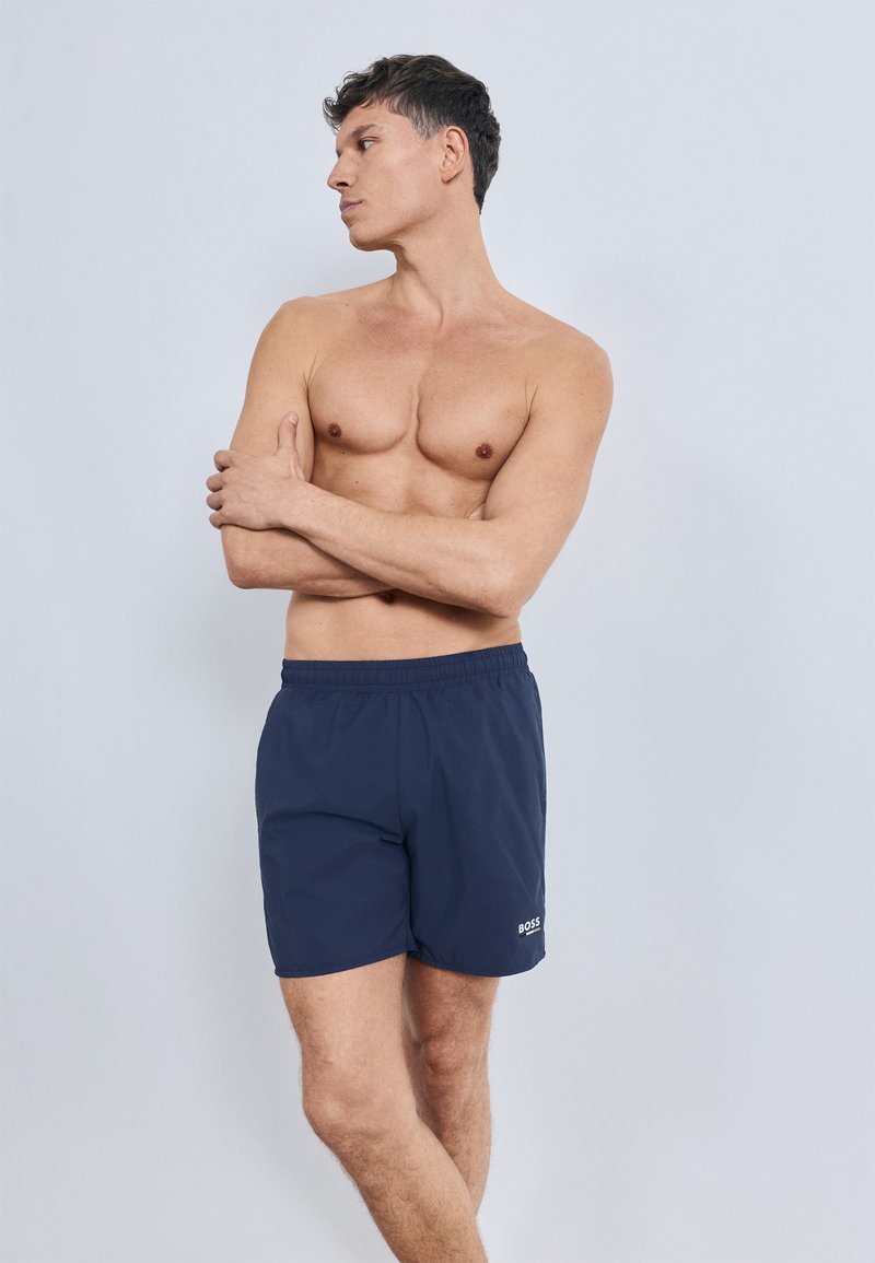 Tengerészkék fürdőnadrág elasztikus derékrésszel és hímzett logóval az alsó lábszáron. Simára szőtt, könnyű anyagból, térd hosszúságú kialakítás.