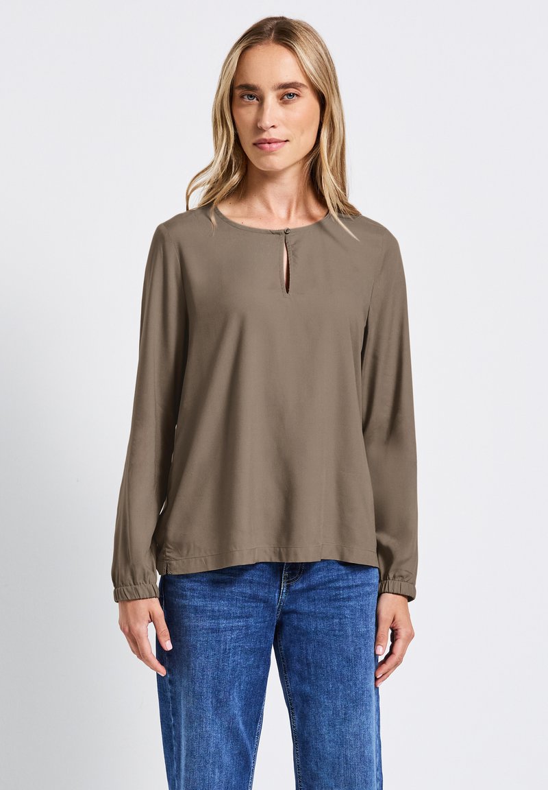 Blusa a maniche lunghe di colore verde oliva con scollatura a chiave e polsini elastici, realizzata in un tessuto morbido, abbinata a jeans blu.