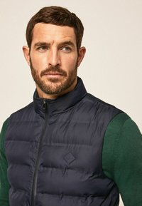 Marineblauer Steppweste mit hochabschließendem Kragen, gestepptem Textil und frontalem Reißverschluss, getragen über einem grünen Pullover. Besitzt ein kleines Logo auf der Brust.