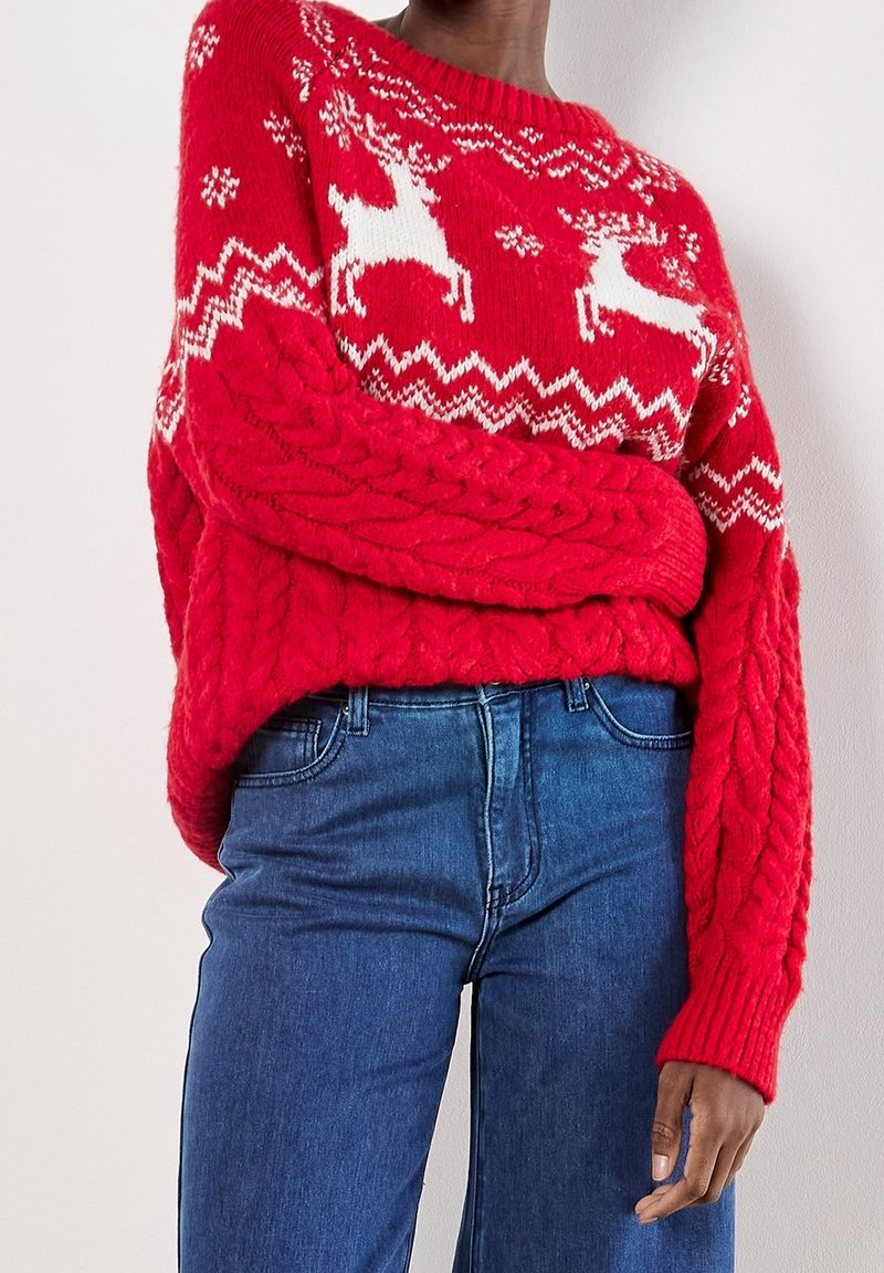 Personne portant un pull rouge en maille torsadée avec des motifs de rennes blancs et de flocons de neige, associé à un jean bleu taille haute.