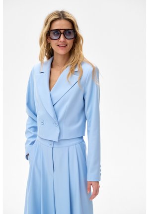 Kobieta w jasnoniebieskim krótkim blezerze i szerokich spodniach, z dużymi ciemnymi okularami przeciwsłonecznymi i kolczykami kołami, uśmiecha się na białym tle.