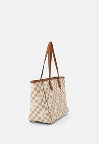 Beige a bílá kostkovaná taška s malými květinovými detaily a hnědými koženými popruhy, stojící vzpřímeně na bílém pozadí.