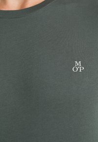 Camiseta verde oscura con cuello redondo y diseño ajustado. Presenta un pequeño logo blanco "M OP" en el lado izquierdo del pecho. Textura suave y lisa.