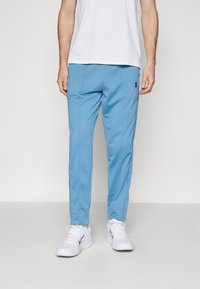 Pantalones deportivos azul claro con textura suave, ajuste entallado y bolsillos laterales, combinados con zapatillas blancas y una camisa blanca lisa.