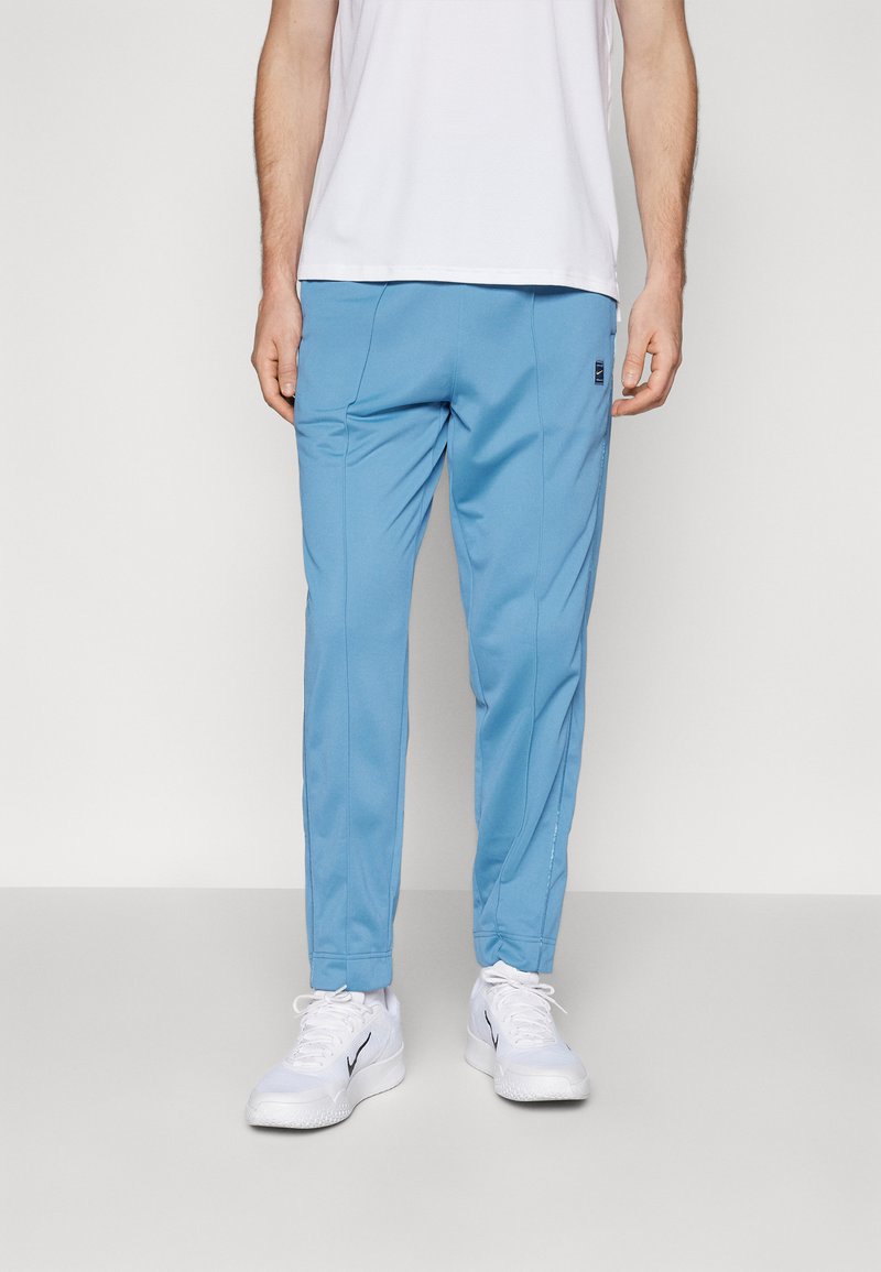Pantalones deportivos azul claro con textura suave, ajuste entallado y bolsillos laterales, combinados con zapatillas blancas y una camisa blanca lisa.