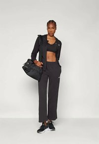 Ženska, oblečena v črno športno modrček, jakno, široke hlače in superge, drži črno športno torbo čez eno ramo pred enobarvno ozadje.