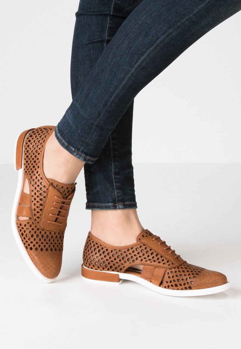 Scarpe in pelle marrone perforata con suole bianche, dotate di un design lace-up e dettagli ritagliati. Indossate con jeans blu scuro.