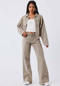 Chaqueta de mezclilla beige con estampado y pantalones acampanados a juego con dobladillos con flecos, llevados sobre una blusa blanca ajustada. Detalles del tejido texturizado visibles.