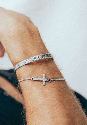 Silver bangle with a textured pattern and a metal chain bracelet featuring a textured cross pendant. Both are worn on a wrist.