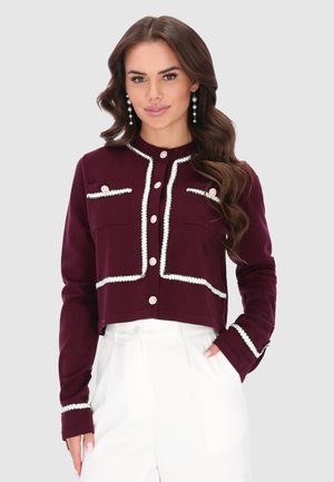 Donna con lunghi capelli mossi, che indossa orecchini di perle, una giacca bordeaux con bordi bianchi e pantaloni bianchi a vita alta, in piedi con una mano in tasca.