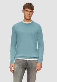 Hellblaues Rundhalsoberteil aus weichem Material, mit gerippten Bündchen und Saum, über einem weißen Hemd getragen, kombiniert mit grauen Jeans.