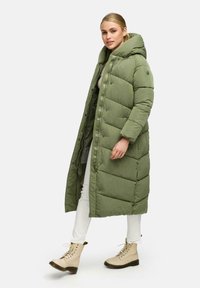 Cappotto lungo e imbottito in verde oliva con cappuccio, caratterizzato da un design trapuntato, una cerniera frontale e chiusure a scatto. Indossato sopra pantaloni bianchi e stivali beige.