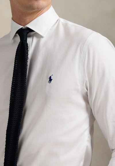White dress shirt with a herringbone texture and a blue embroidered logo. Paired with a dark knitted tie, featuring a textured design.