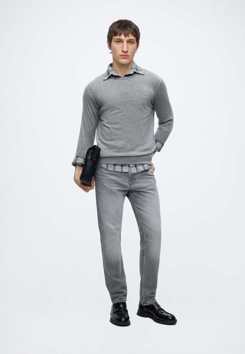 Young man wearing a gray sweater, plaid shirt, and gray jeans stands holding a black leather clutch against a plain white background.