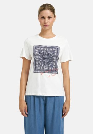 Frau trägt ein weißes T-Shirt mit einem dunkelblauen, filigranen Quadratmuster und rotem kursivem Schriftzug, kombiniert mit locker sitzenden blauen Hosen, steht vor einem einfarbigen Hintergrund.