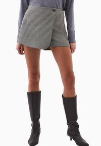 Kvinde iført ternet omslag-mini nederdel, grå langærmet top og sorte knæhøje hæle støvler, stående foran en hvid baggrund.