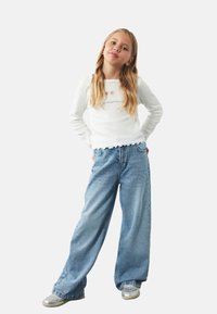 Witte lange mouwen top met gegolfde randen en decoratieve uitsparingen, gecombineerd met lichtblauwe wijde jeans met strass-accenten. Zilveren schoenen.