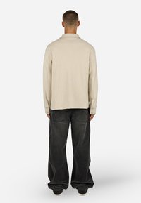 Beige hosszúujjú ing, texturált anyagból, gallérral és gombos mandzsettával, laza szabású fekete farmernadrággal kombinálva.