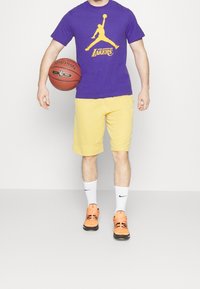 Fialové tričko s žlutým grafickým logem, žluté šortky, bílé ponožky a oranžovo-černé sportovní boty, držící basketbal.