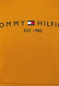 Maglione in cotone arancione con ricamo nero della scritta 'TOMMY HILFIGER', corredato da un logo rettangolare in rosso, bianco e blu. Fondato nel 1985.