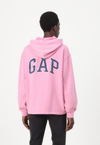 Sweat-shirt à capuche en tissu rose clair, arborant un grand logo "GAP" navy au dos, avec des manches longues et une coupe décontractée.