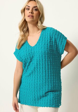 Femme blonde portant un haut turquoise texturé à manches courtes et col en V, debout devant un fond clair uni.