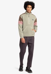 Fleece pullover in muted green with brown, pink, and cream color-blocked sleeves. Features a half-zip collar and a front pocket.