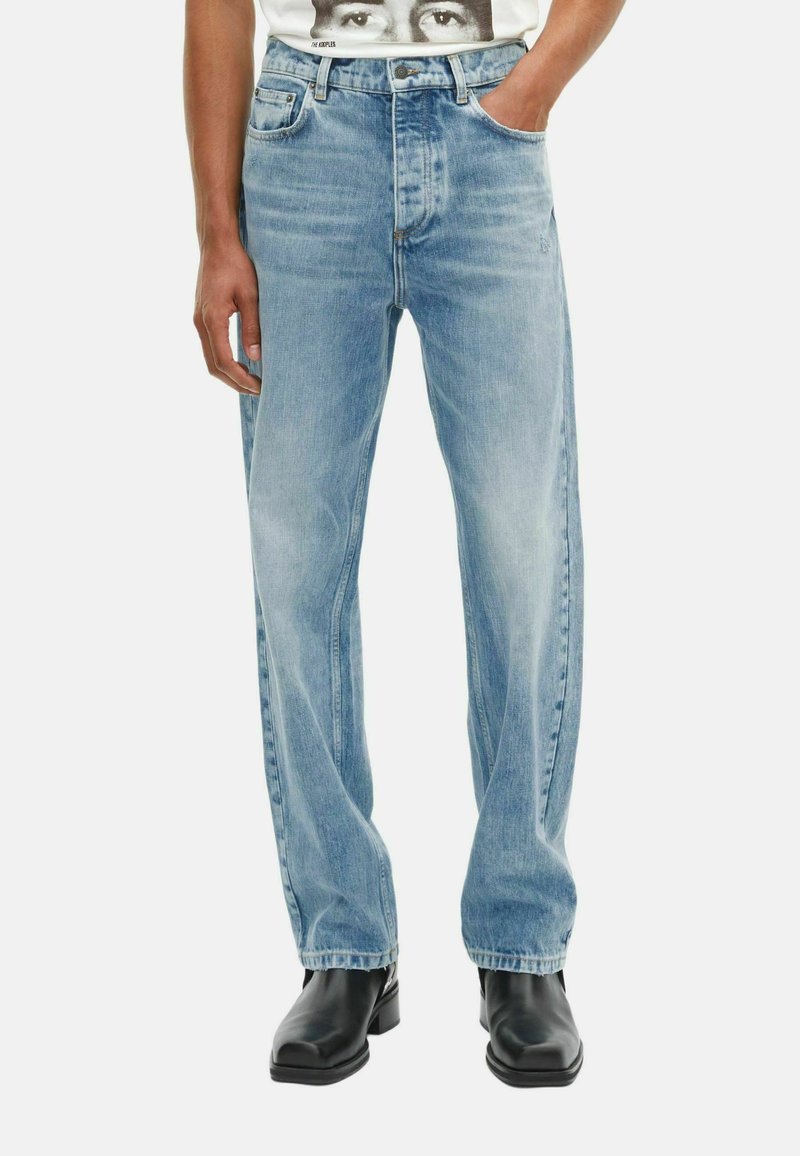 Persoon in lichtblauwe rechte jeans met handen in de zakken en zwarte vierkante laarzen op een witte achtergrond.