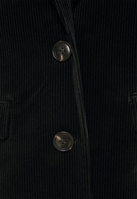 Černé kordové sako s vertikálním žebrováním, dvěma černými knoflíky a bočními kapsami. Texturovaná látka s hladkým povrchem.