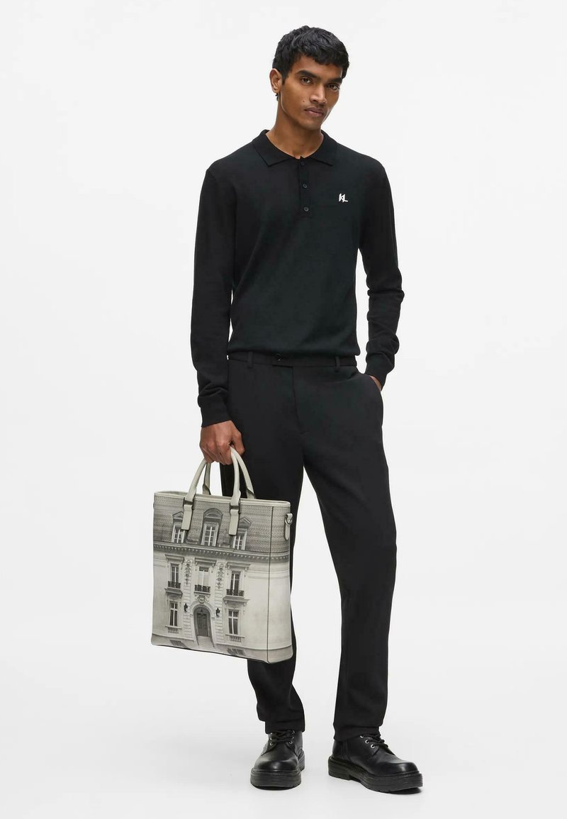 Homme portant un polo noir à manches longues et un pantalon, tenant un sac cabas beige avec un imprimé bâtiment en noir et blanc, debout devant un fond blanc uni.