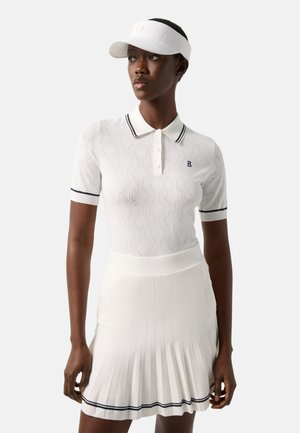 Kvinde iført en hvid tekstureret polo i sort kant, hvid plisseret nederdel med sorte striber og hvid skygge, stående foran en ensfarvet baggrund.