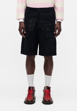 Person iført sorte cargoshorts med et subtilt dyremønster, hvide crew-strømper, brune støvler med klare røde snørebånd og en pink- og hvidstribet langærmet skjorte.