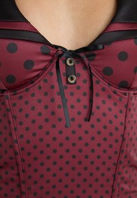Burgundy corset featuring black polka dots, satin trim, a V-neckline, and a decorative bow with button accents at the center.