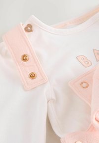 Valkoinen vauvan paita, jossa on vaaleanpunaiset olkaimet ja kultaiset painonapit sekä hopealla kirjailtu teksti "BABY". Pehmeä puuvillamateriaali, jolla on sileä rakenne.