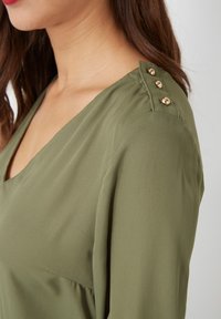 Mujer con blusa verde oliva de escote en V y tres botones dorados en la costura del hombro, mostrando el perfil lateral y cabello castaño.