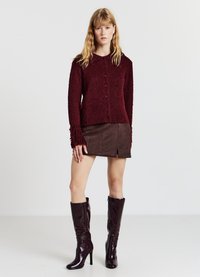 Bordeauxfarbener strukturierter Cardigan mit Kragen, vorderen Knöpfen und Fransen an den Ärmeln, kombiniert mit einem braunen Wildleder-Minirock und kniehohen Stiefeln.